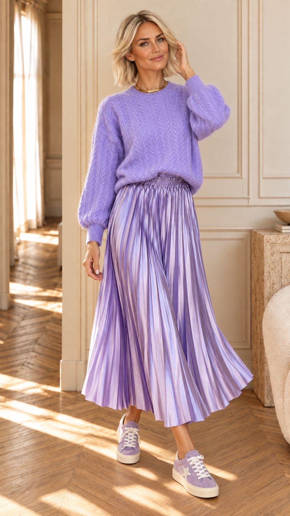 A woman stands in a sunlit interior wearing a lavender knit sweater and a matching pleated midi skirt with a smocked waist, paired with light purple sneakers.