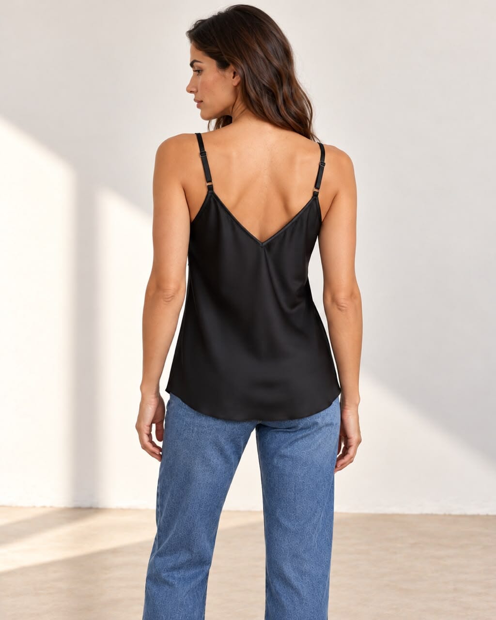 A woman modeling a black satin camisole top with black lace trim along the V-neckline, paired with blue jeans, standing against a light beige wall with soft shadows.